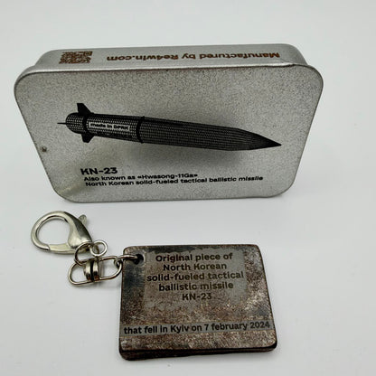 Rectangular KN-23 missile fragment keyring inscribed with Kyiv, 7 February 2024, beside a RE4WIN presentation tin showing the missile.