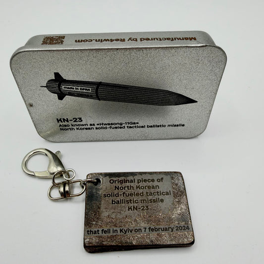 Rectangular KN-23 missile fragment keyring inscribed with Kyiv, 7 February 2024, beside a RE4WIN presentation tin showing the missile.