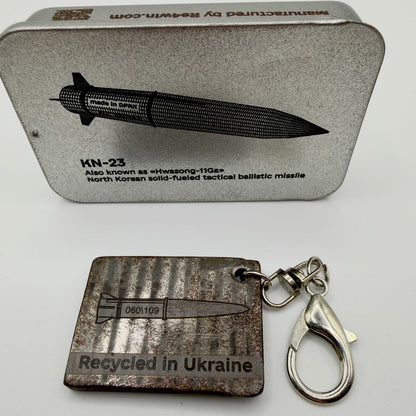 Dark metal fragment keyring with a missile illustration and “Recycled in Ukraine” inscription, attached to a silver-tone swivel and lobster clasp.