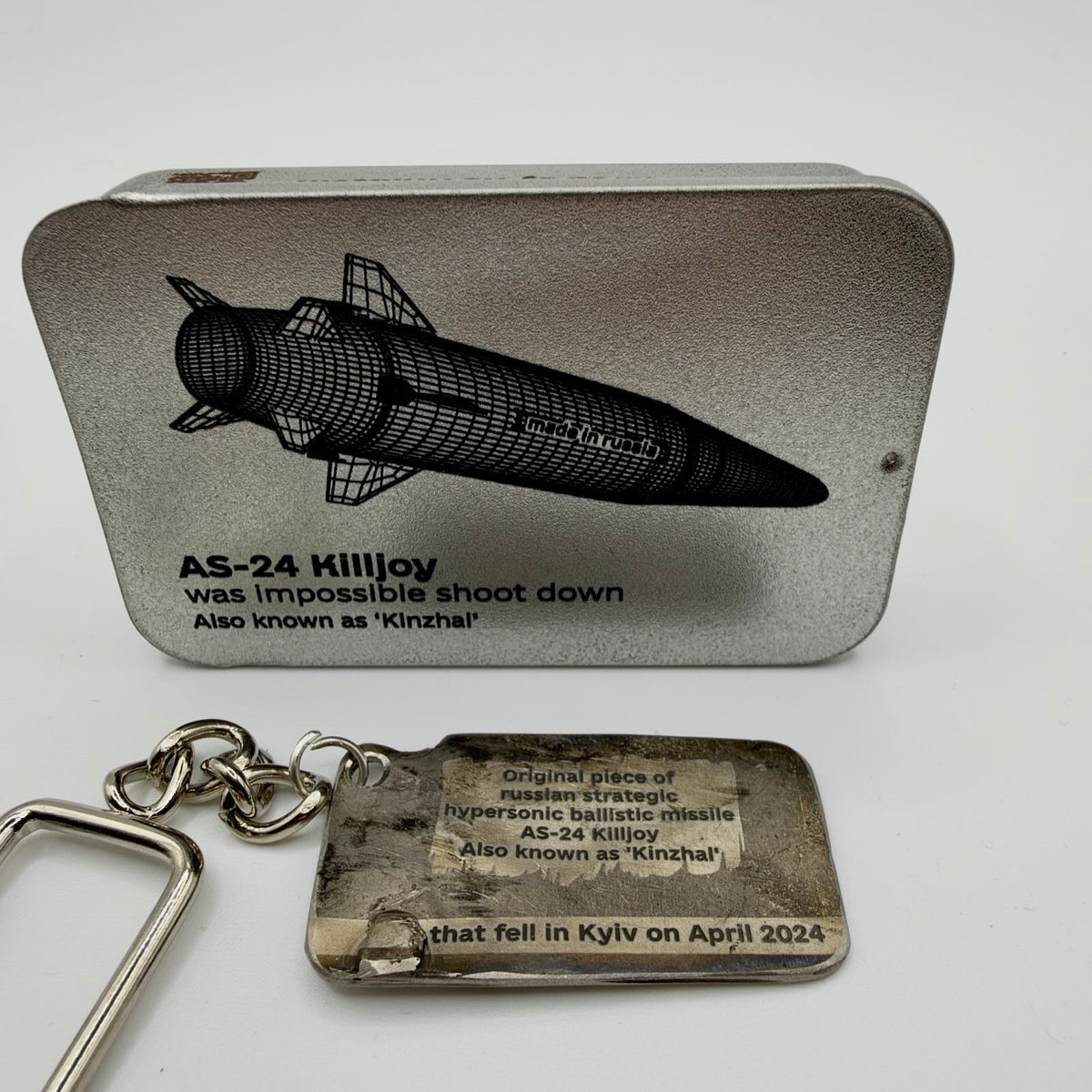 Missile fragment keyring inscribed as a Kinzhal AS-24 piece that fell in Kyiv in April 2024, pictured beside an illustrated presentation tin.