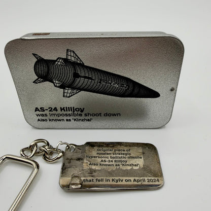 Missile fragment keyring inscribed as a Kinzhal AS-24 piece that fell in Kyiv in April 2024, pictured beside an illustrated presentation tin.
