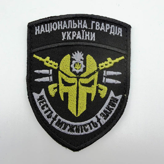 Black shield-shaped embroidered patch with the Ukrainian National Guard’s name, a yellow skull-style face beneath a small Guard emblem, crossed rifles, grey cartridge motifs and a banner reading “Honour, Courage, Law.”