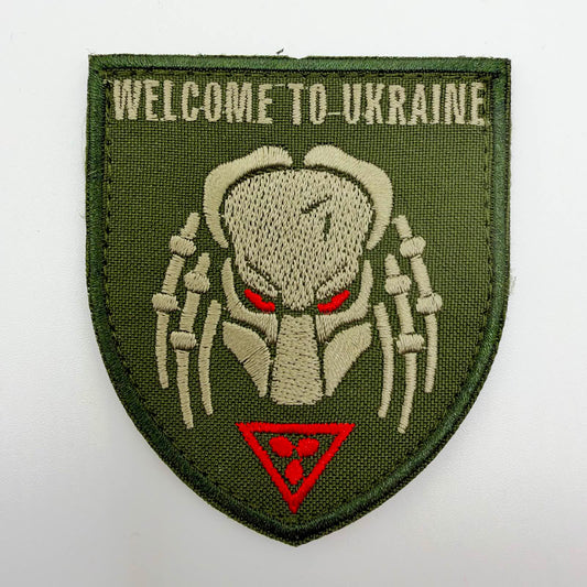 Olive-green shield-shaped embroidered patch with “WELCOME TO UKRAINE” above a khaki Predator-style mask with red eyes, and a red inverted triangle containing three red dots below.