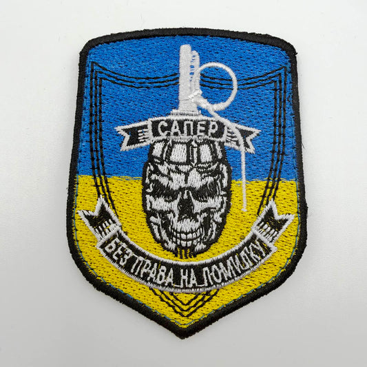 Blue-and-yellow shield-shaped embroidered patch with black edging, a white-and-black skull incorporated into a grenade design, and Ukrainian banners reading “Sapper” and “No Room for Error.”