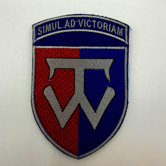 Shield-shaped embroidered 58th Brigade patch with red and blue halves, a large silver-grey central emblem and border, and the motto “SIMUL AD VICTORIAM” across the blue top panel.