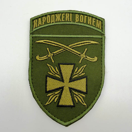 Olive-green shield-shaped patch with the Ukrainian words “Born of Fire” above crossed khaki sabres and a khaki cross surrounded by stylised flames.