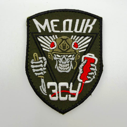 Olive shield-shaped embroidered medic morale patch with a black border, white skull wearing a winged helmet with red crosses, skeletal hands holding medical equipment, and white Ukrainian “МЕДИК” and “ЗСУ” lettering.