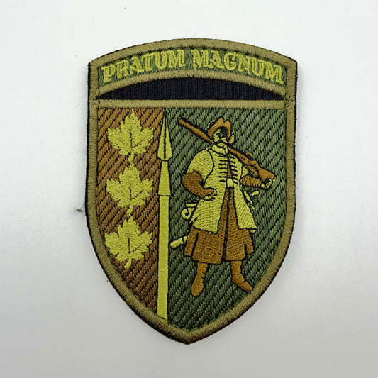 Shield-shaped embroidered patch in olive, brown and gold tones, featuring a Cossack carrying a musket, a vertical spear, three maple leaves and the Latin inscription “PRATUM MAGNUM”