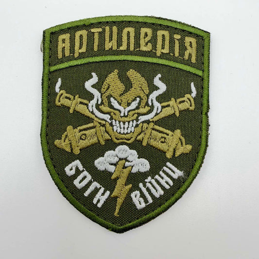 Olive shield-shaped embroidered artillery morale patch with green edging, a gold-toned skull over crossed cannon barrels, white smoke, a cloud and lightning bolt, and Ukrainian lettering meaning “Artillery” and “Gods of War”.