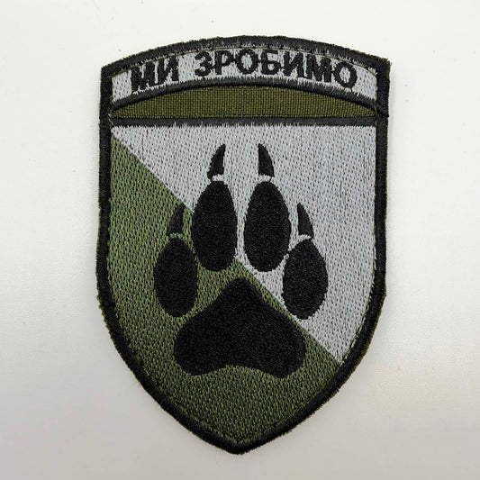 Shield-shaped embroidered patch with a black border, diagonally divided olive-and-light-grey background, a large black paw print with claws and Ukrainian “МИ ЗРОБИМО” lettering across the top.