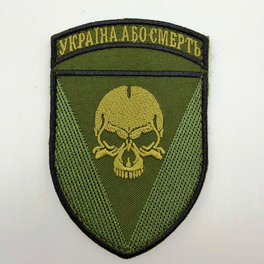 Olive shield-shaped embroidered patch with a black border, central gold-toned skull, contrasting green triangular panels and Ukrainian “Україна або смерть” lettering across the top.