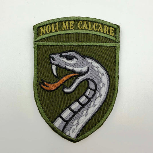 Olive shield-shaped embroidered patch featuring a grey-and-white viper with exposed fangs and an orange forked tongue, beneath the gold Latin motto “NOLI ME CALCARE”.