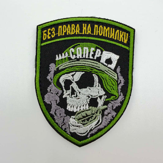 Black shield-shaped embroidered sapper morale patch with bright green edging, a white skull wearing a green helmet and biting a grenade, yellow Ukrainian “No room for error” lettering and white “Sapper” text.


16:07