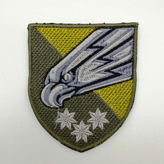 Olive shield-shaped embroidered patch with a large silver-grey falcon’s head angled downwards, yellow triangular accents and three silver-grey stars beneath.