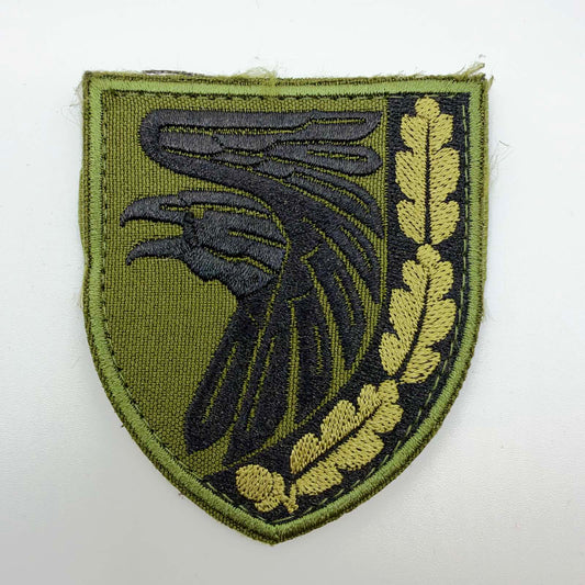 Olive-green shield-shaped embroidered patch featuring a large black raven and a golden-olive oak branch with leaves and an acorn along the right edge, without lettering.