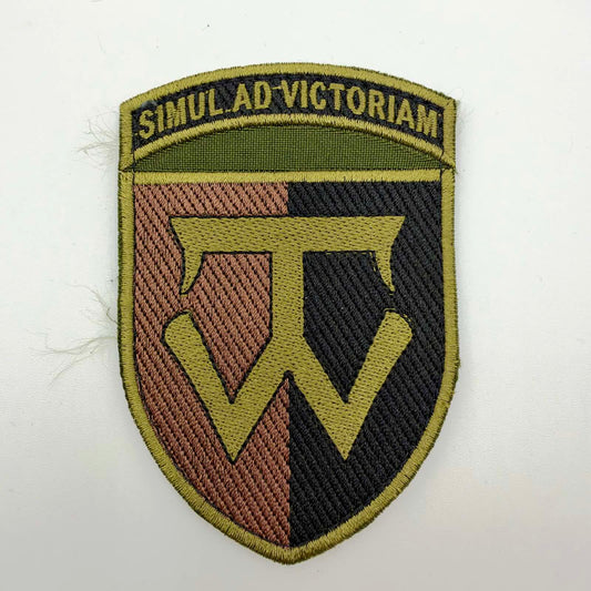 Shield-shaped embroidered patch with a divided brown-and-black background, an olive-coloured central emblem and border, and the Latin motto “SIMUL AD VICTORIAM” across the top.