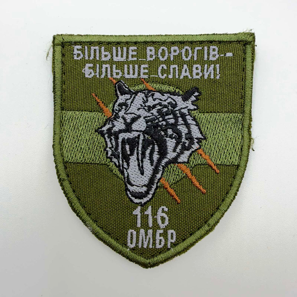 Olive-green shield-shaped embroidered patch featuring a snarling white-and-black tiger, orange claw marks, “116 ОМБр” below, and the Ukrainian slogan “More enemies — more glory” above.