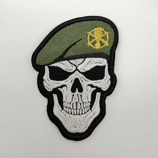 Skull-shaped embroidered morale patch featuring a white grinning skull with black eye sockets, wearing an olive-green beret with a gold-coloured emblem and black edging.