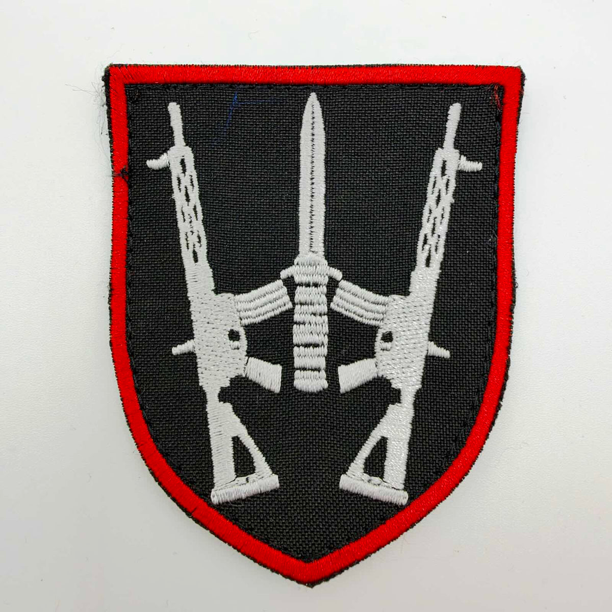 Black shield-shaped embroidered patch with a red border and two white rifles flanking a central upright blade, arranged in a stylised trident.