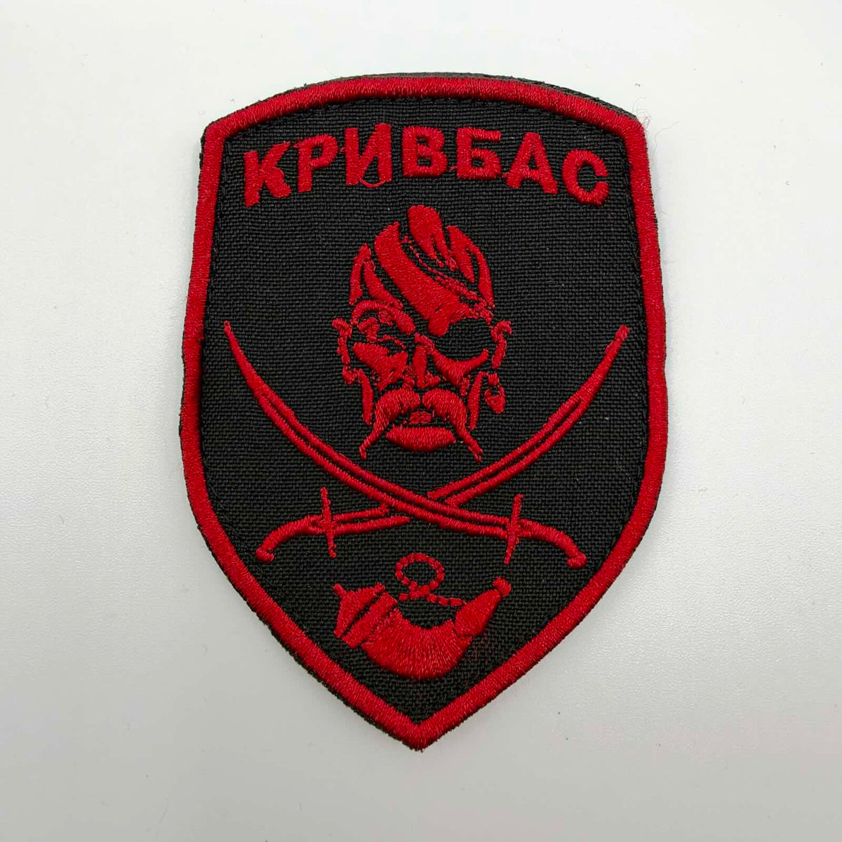 Black shield-shaped patch with red embroidery reading “Kryvbas”, featuring a moustached Cossack wearing an eye patch, crossed sabres and a curved horn.