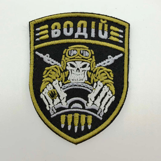 Black shield-shaped embroidered morale patch with olive-yellow edging, Ukrainian text meaning “Driver”, and a skull-faced driver wearing goggles and gripping a steering wheel, with crossed rifles and cartridge motifs.