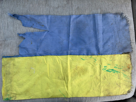 Worn Ukrainian flag laid flat on a grey sofa, blue above yellow, with large tears and missing sections in the blue fabric, frayed edges, staining and green marks.