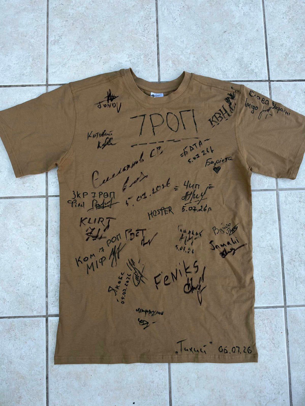 Front of a khaki military T-shirt covered in handwritten signatures, callsigns and dates, with large “7 РОП” lettering across the chest and “Glory to Ukraine” in Ukrainian on one sleeve.