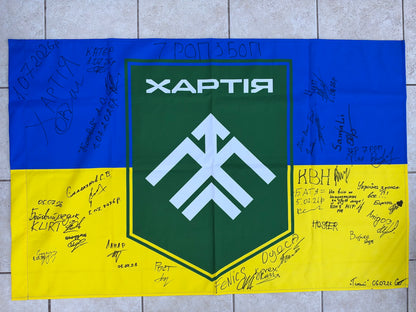 Blue-and-yellow Ukrainian flag with a large green Khartiia shield and white emblem, surrounded by handwritten signatures, messages and July 2026 dates.