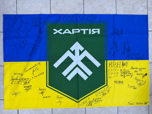 Blue-and-yellow Ukrainian flag with a large green Khartiia shield and white emblem, surrounded by handwritten signatures, messages and July 2026 dates.
