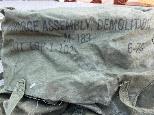 Close-up of the bag’s faded black M-183 demolition assembly markings, dusty fabric and frayed flap edge.