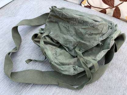 Heavily worn olive-green military shoulder bag with webbing straps and a flap marked “Charge Assembly, Demolition M-183”.