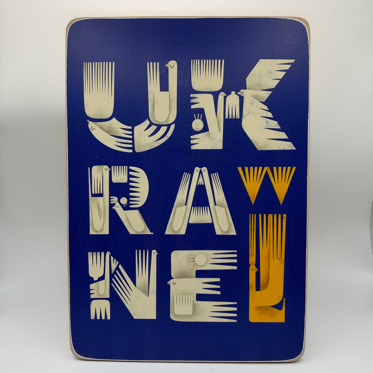 Blue wooden wall plaque spelling “Ukraine” in cream stylised bird lettering, with yellow bird motifs and rounded corners.