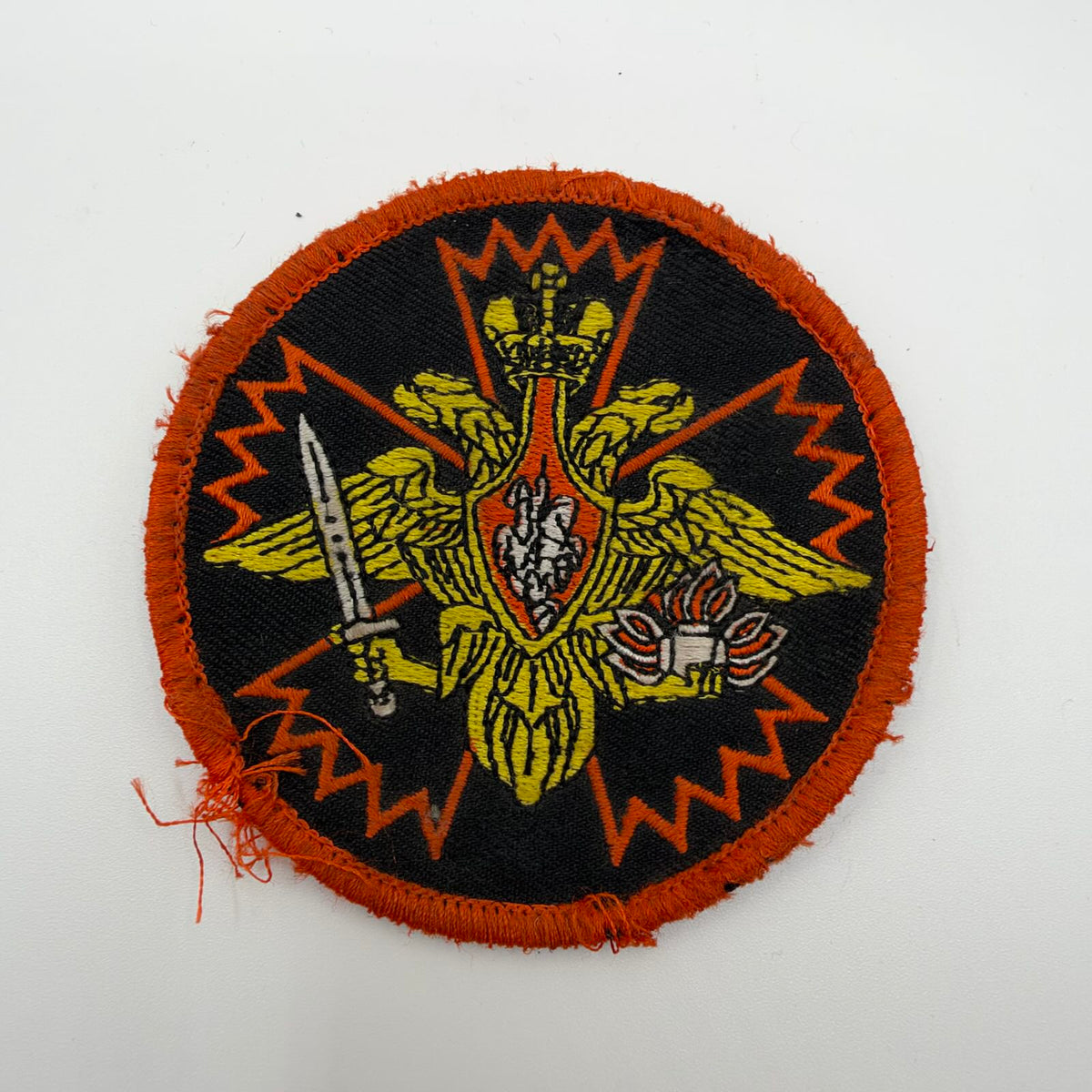 IMG_0602.jpeg: Worn circular Russian patch with a gold double-headed eagle, sword and flaming grenade over an orange carnation outline, with a frayed orange border.
