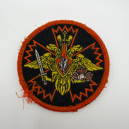 IMG_0602.jpeg: Worn circular Russian patch with a gold double-headed eagle, sword and flaming grenade over an orange carnation outline, with a frayed orange border.