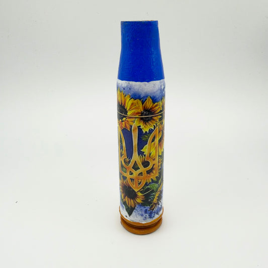 Decorated shell casing with a blue neck and golden-brown base, featuring a large gold Ukrainian trident surrounded by yellow sunflowers and green leaves against blue and white.
