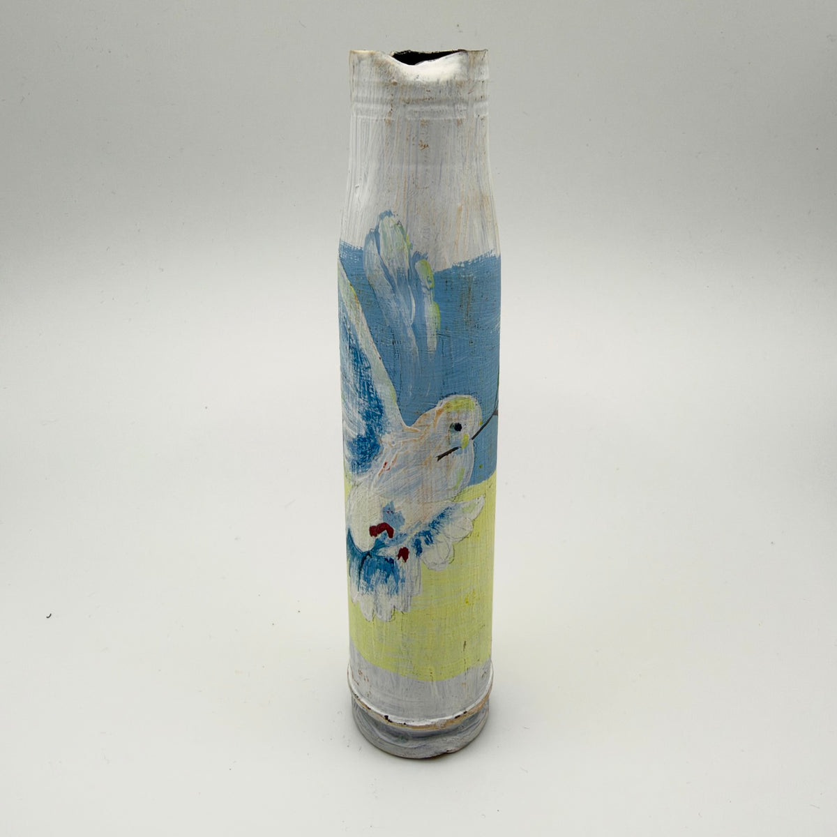 White-painted shell casing with a dented opening, featuring a white dove with blue feather details and outstretched wings over pale blue and yellow bands.