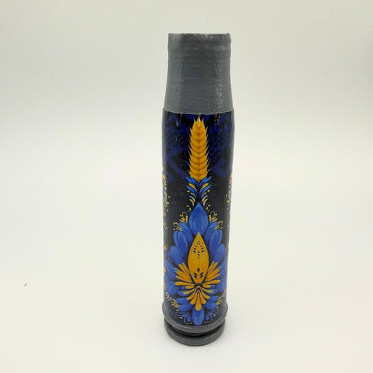Decorated shell casing with a grey neck and base, featuring a golden ear of wheat above an ornate blue and yellow floral design on a dark background.