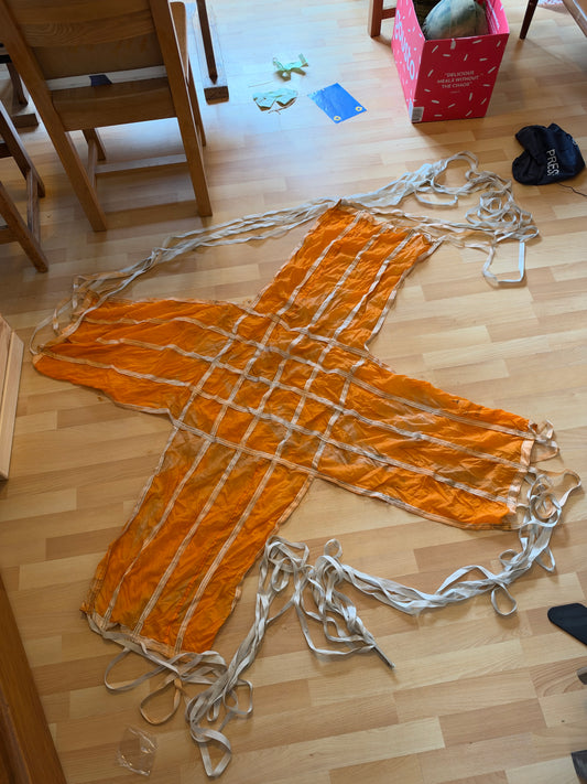Orange cross-shaped parachute canopy spread flat on a wooden floor, with a grid of white reinforcing tapes and long white suspension tapes extending from its four arms.