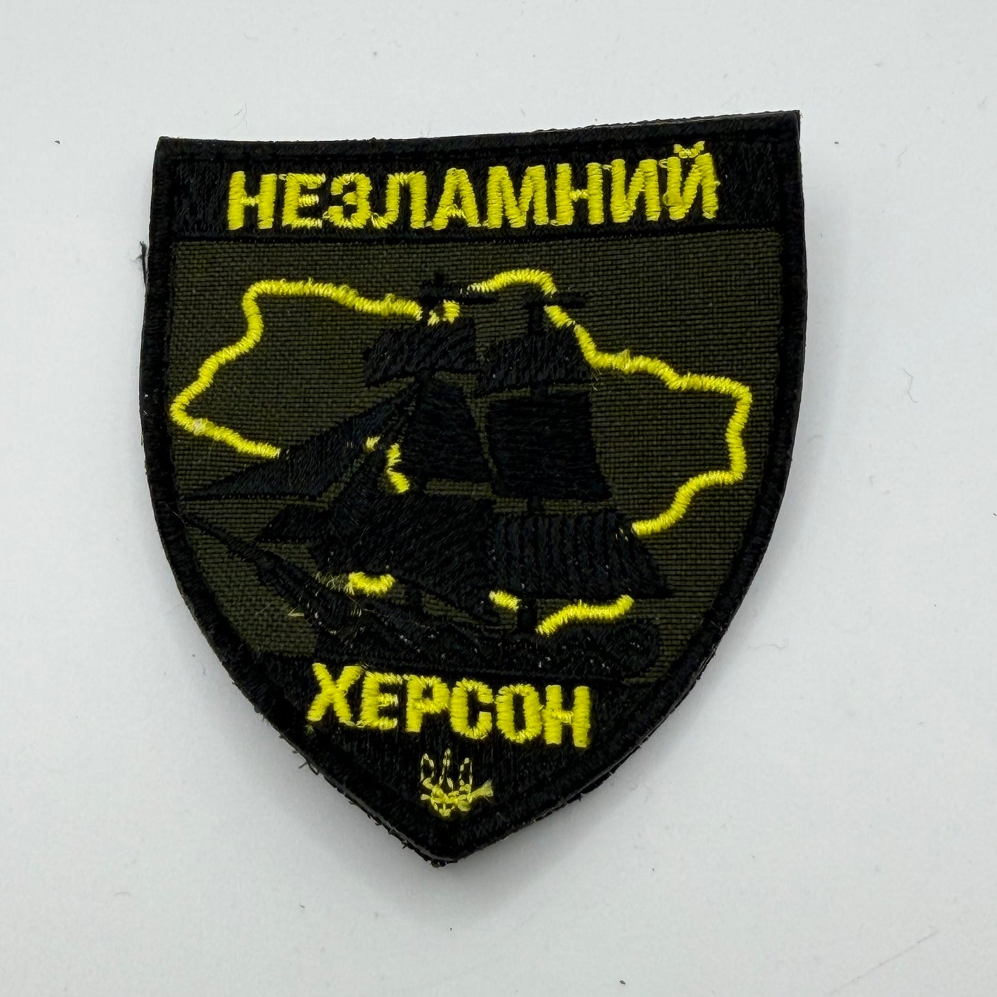 Black and gold patch reading Unbreakable Kherson, and depicting on outline of Kherson Oblast and showing a sailing ship