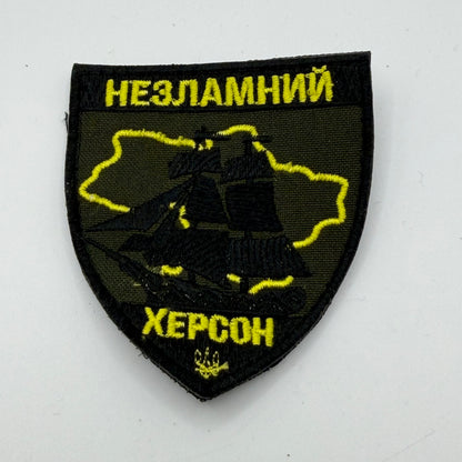 Black and gold patch reading Unbreakable Kherson, and depicting on outline of Kherson Oblast and showing a sailing ship
