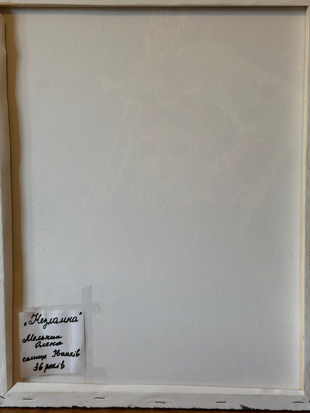 Reverse of the canvas showing its wooden stretcher frame and a handwritten Ukrainian label in the lower-left corner. The label reads “Unbreakable”, Olena Melnyk, Ivankiv, aged 36.
