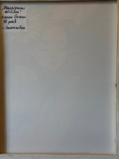 Reverse of the canvas showing its wooden stretcher frame and a handwritten Ukrainian label in the upper-left corner. The label gives the title “The Dearest Embrace” and identifies the artist as Oksana Karas, aged 46, from Stanishivka village.