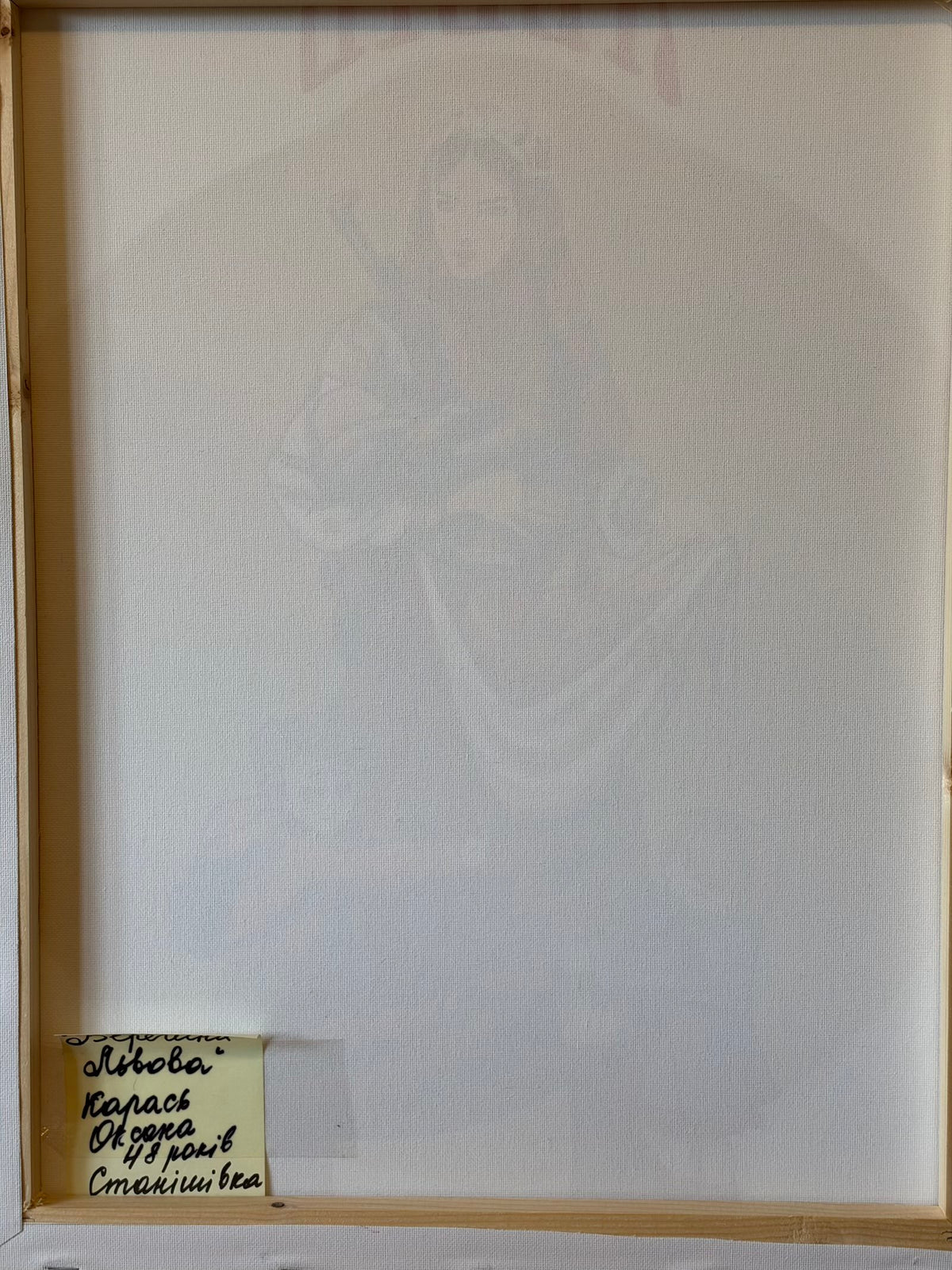 Reverse of the canvas showing its wooden stretcher frame and a folded handwritten Ukrainian label at the lower left. It identifies Oksana Karas, aged 48, from Stanishivka. The partly concealed title appears to read “Guardian of Lviv”.