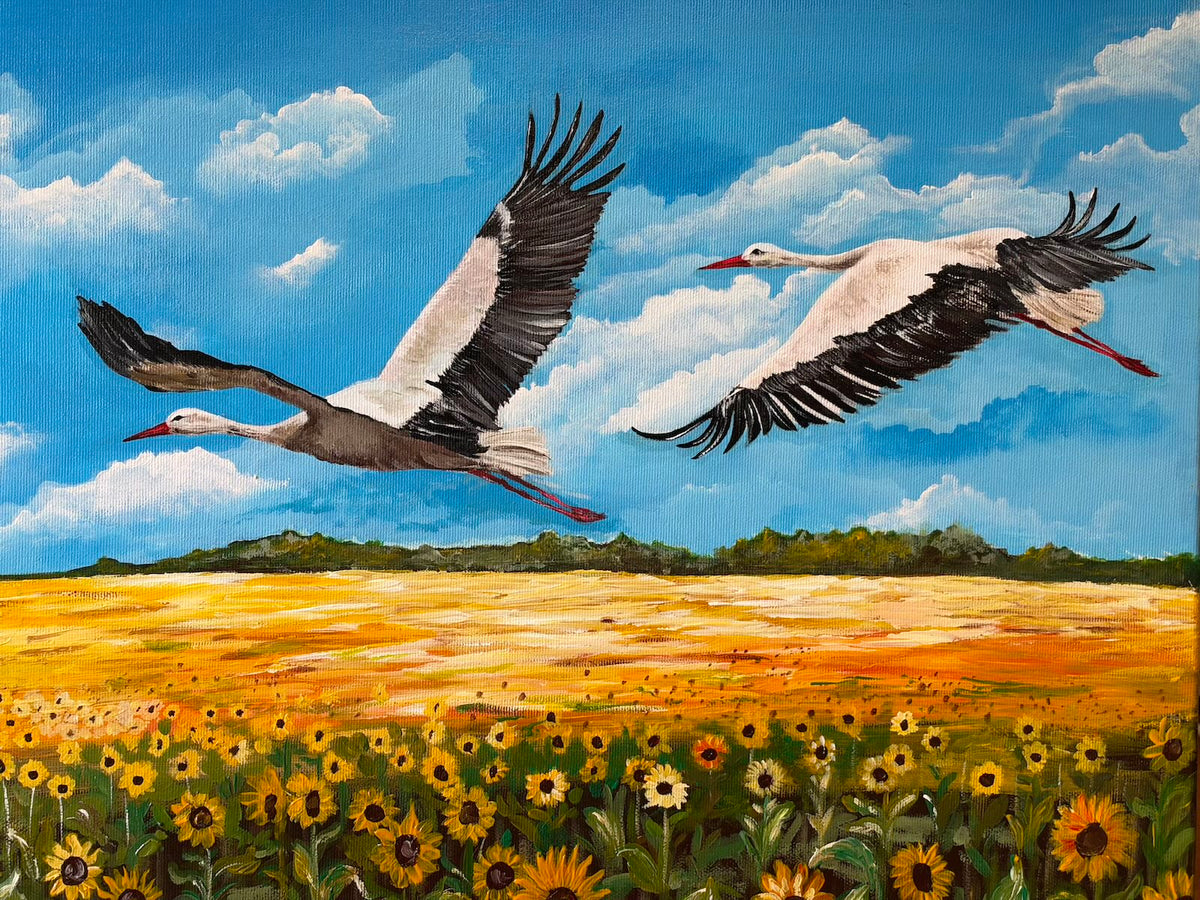 “Storks”, an oil painting of two white storks with black wing feathers, red beaks and trailing red legs flying above golden fields. Sunflowers fill the foreground beneath a bright blue sky scattered with white clouds and a distant green tree line.