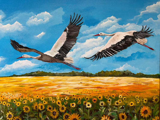 “Storks”, an oil painting of two white storks with black wing feathers, red beaks and trailing red legs flying above golden fields. Sunflowers fill the foreground beneath a bright blue sky scattered with white clouds and a distant green tree line.