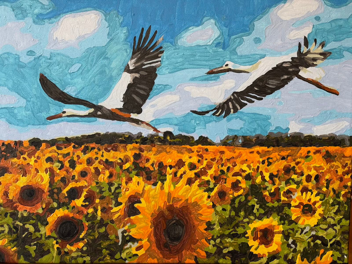 “Sunflowers and Storks”, an oil painting by Olena showing two black-and-white storks flying above a dense field of golden-yellow and orange sunflowers. Green foliage and a distant dark tree line sit beneath a turquoise-blue sky with sweeping white clouds.