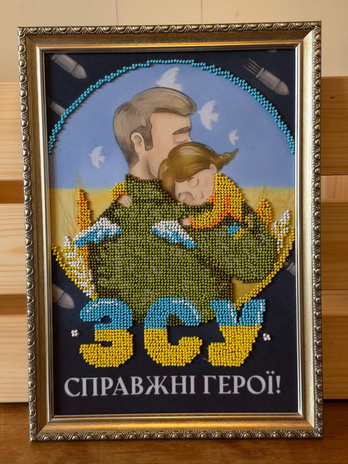 Framed beadwork picture of a Ukrainian soldier embracing a child. Green beads form a map of Ukraine across the central figures, surrounded by beaded wheat, white doves and blue-and-white ribbons. Blue-and-yellow letters read “ЗСУ”, above Ukrainian text meaning “True Heroes”. An ornate metallic-look frame surrounds the picture.