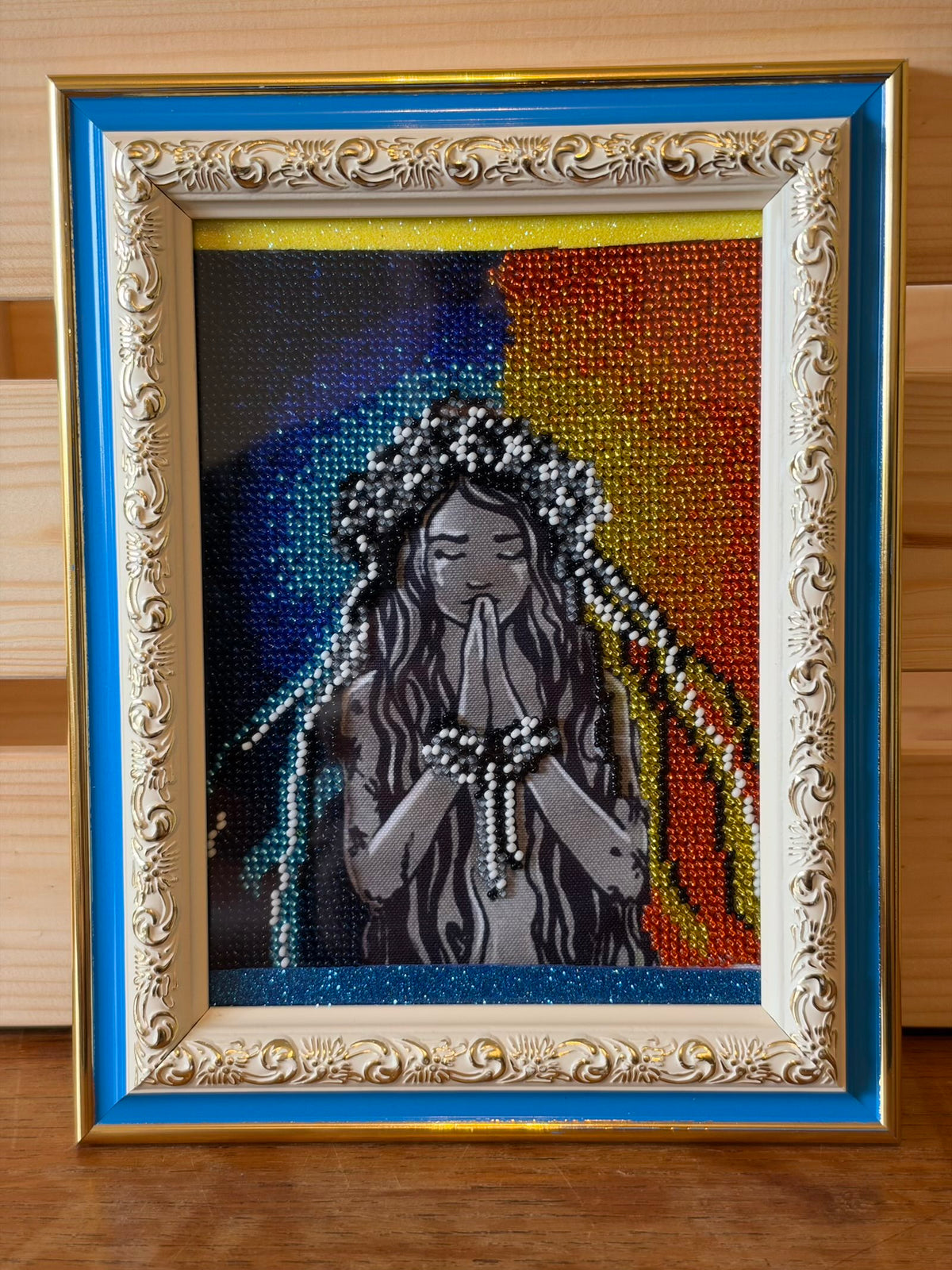 Framed beadwork picture of a long-haired woman wearing a floral wreath, eyes closed and hands together in prayer. White and dark beads decorate her wreath, ribbons and wrists, surrounded by blue, turquoise, gold and orange beadwork. The ornate frame is cream and gold with a bright blue outer border.