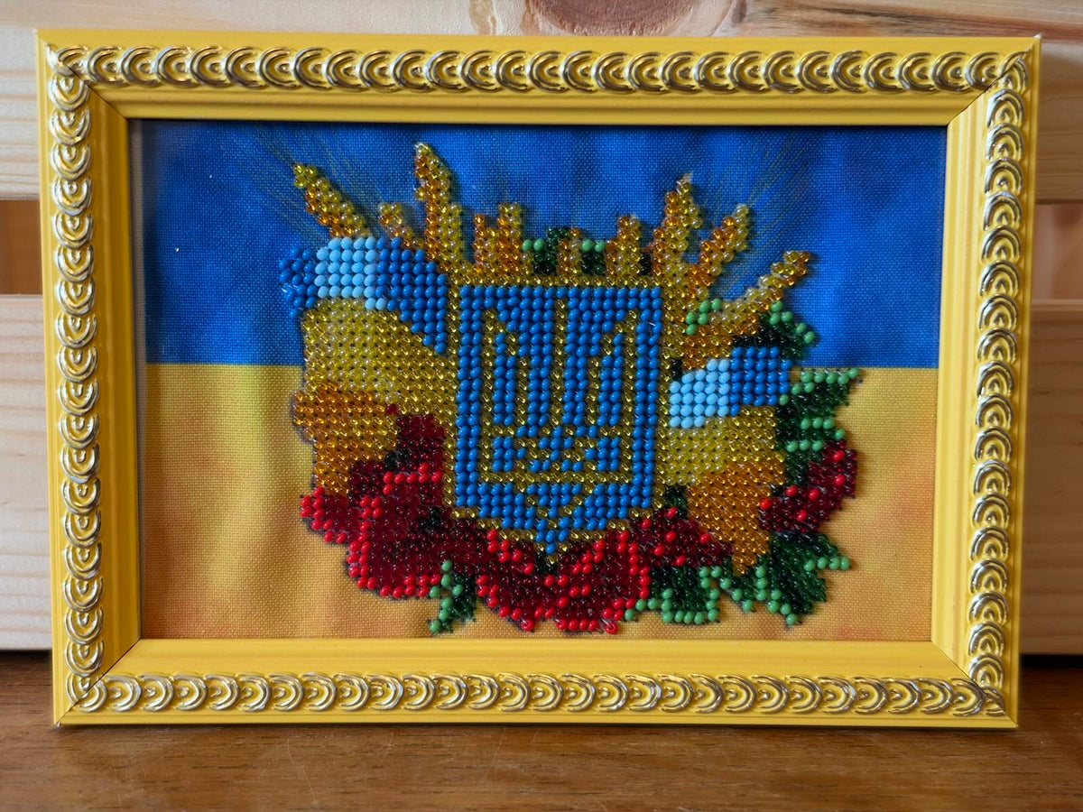 Small framed beadwork depicting Ukraine’s trident coat of arms in blue and gold, surrounded by golden wheat, red poppies, green leaves and a blue ribbon. The background is blue above yellow, and the decorative frame is gold-coloured.