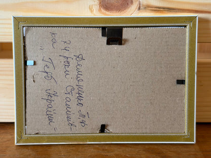 Reverse of the framed beadwork showing a brown backing board, retaining clips and a hanging fitting. The handwritten Ukrainian inscription identifies Tamara Demianenko, aged 74, Stanishivka, and the title “Coat of Arms of Ukraine”.